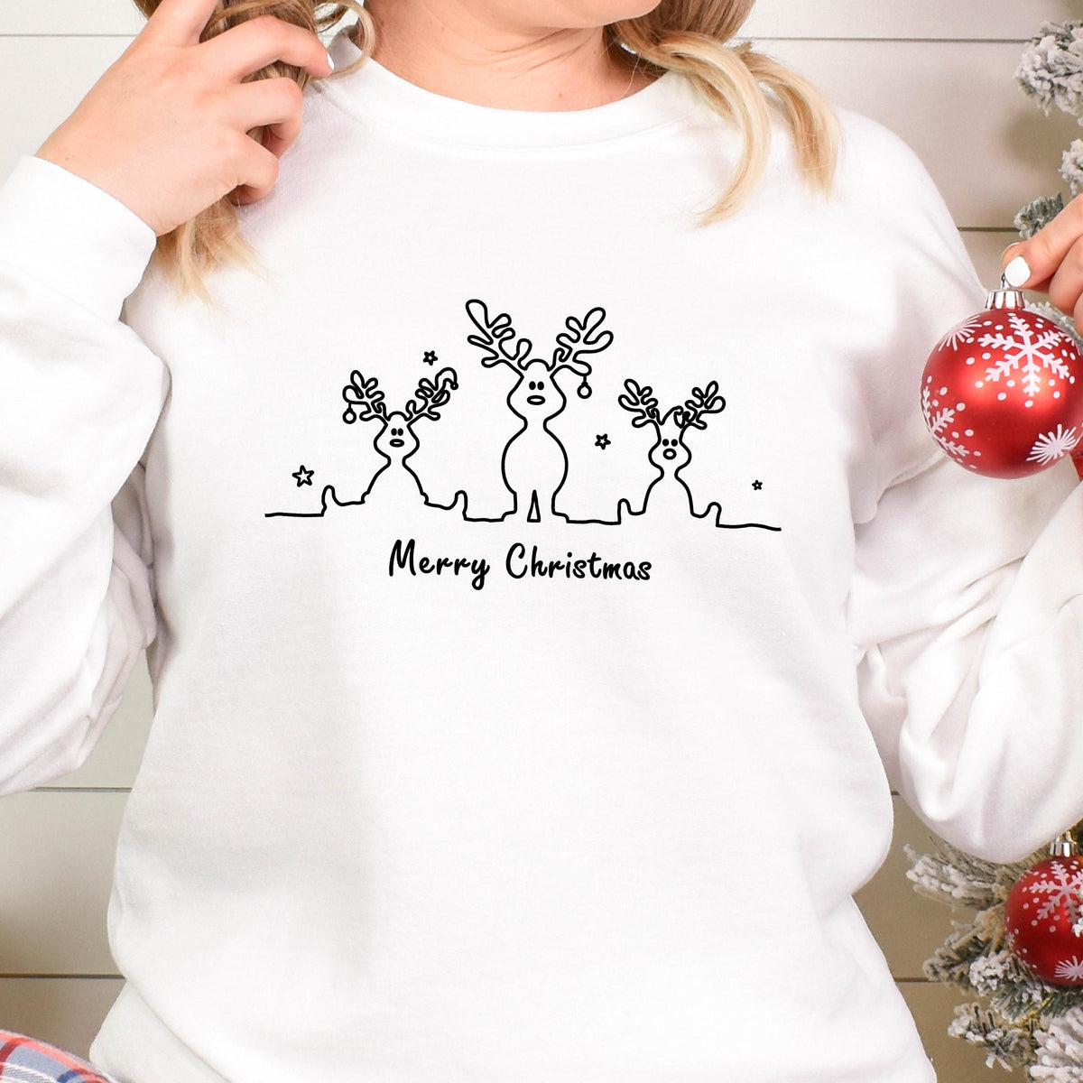 May include: A white long-sleeve sweatshirt featuring a black line art graphic of three reindeer with antlers and small stars. Below the reindeer, the text Merry Christmas is printed in a simple black font. The design is centered on the chest.