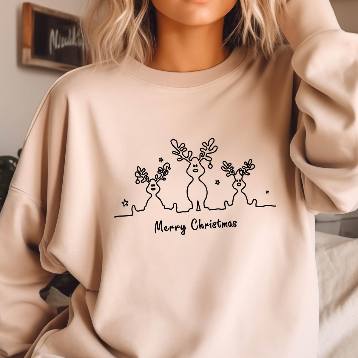 May include: A beige crewneck sweatshirt featuring a black line art graphic of three reindeer with small stars and the text Merry Christmas printed below the figures. The design is centered on the chest of the long-sleeved pullover garment.