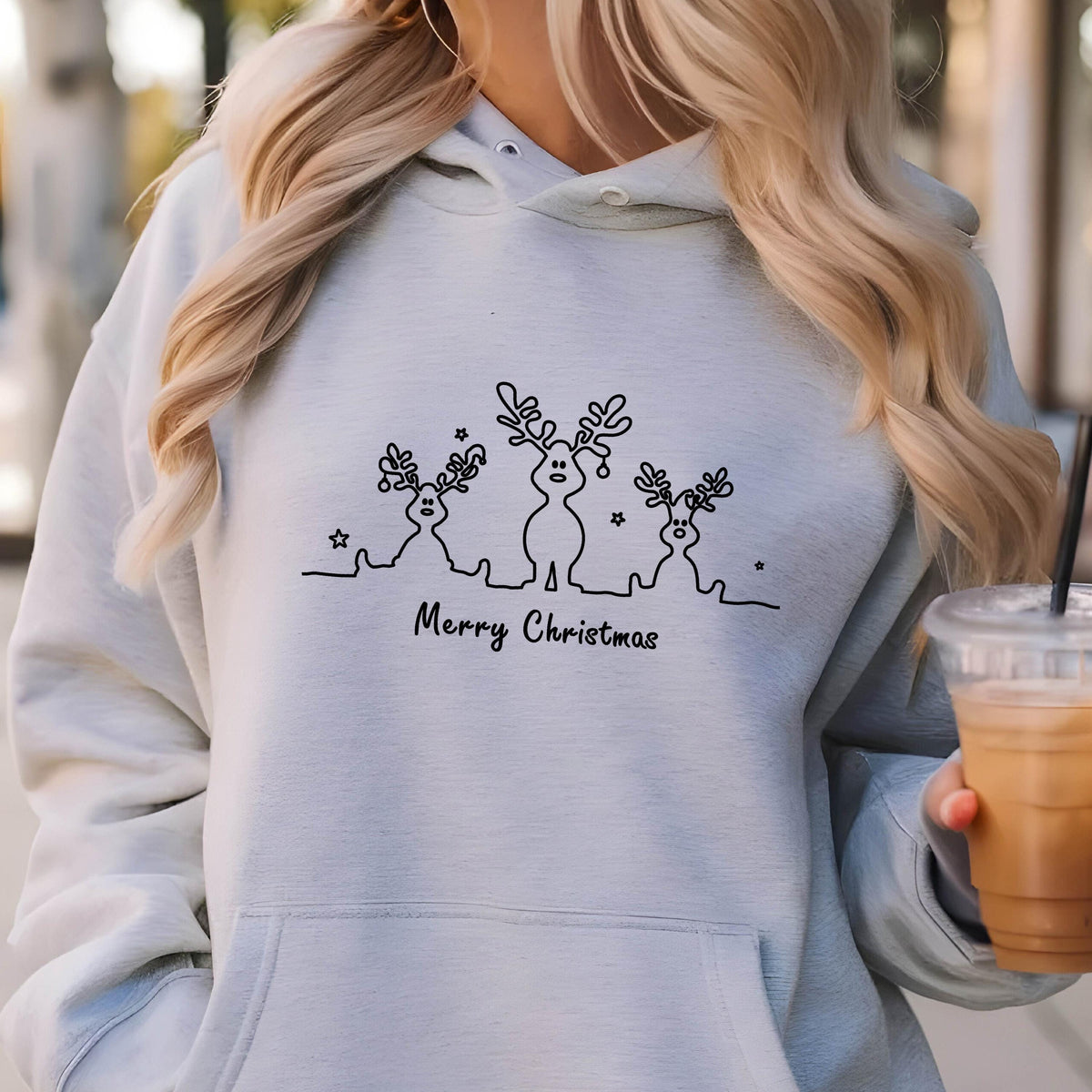 May include: A light blue hooded sweatshirt featuring a black line art graphic of three reindeer with antlers and small stars. The text Merry Christmas is printed in black script below the reindeer figures. The garment has a front kangaroo pocket.