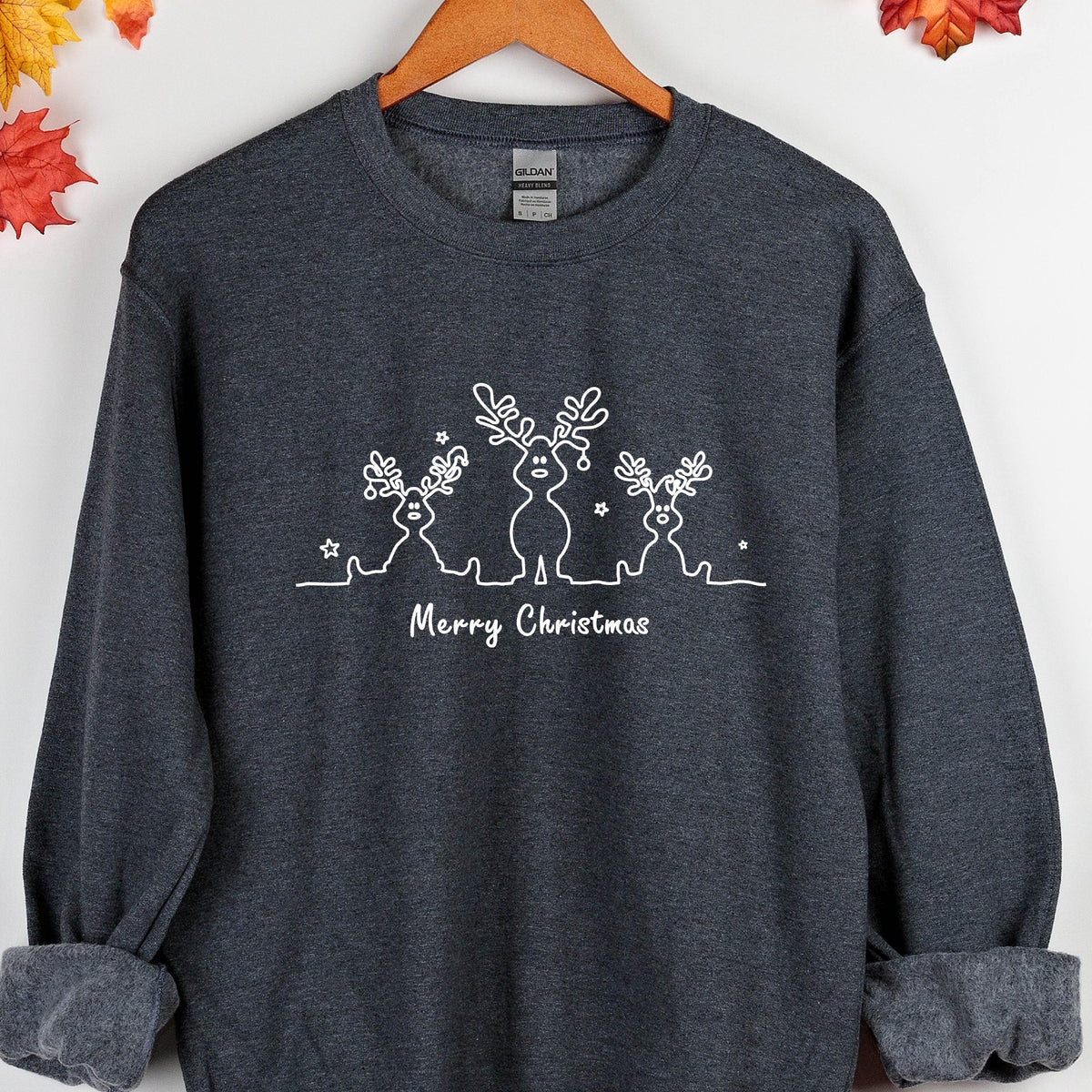 May include: A dark heather gray crewneck sweatshirt featuring a white line art graphic of three reindeer and small stars. Below the reindeer, the text Merry Christmas is printed in a white script font. The garment has long sleeves and ribbed cuffs.