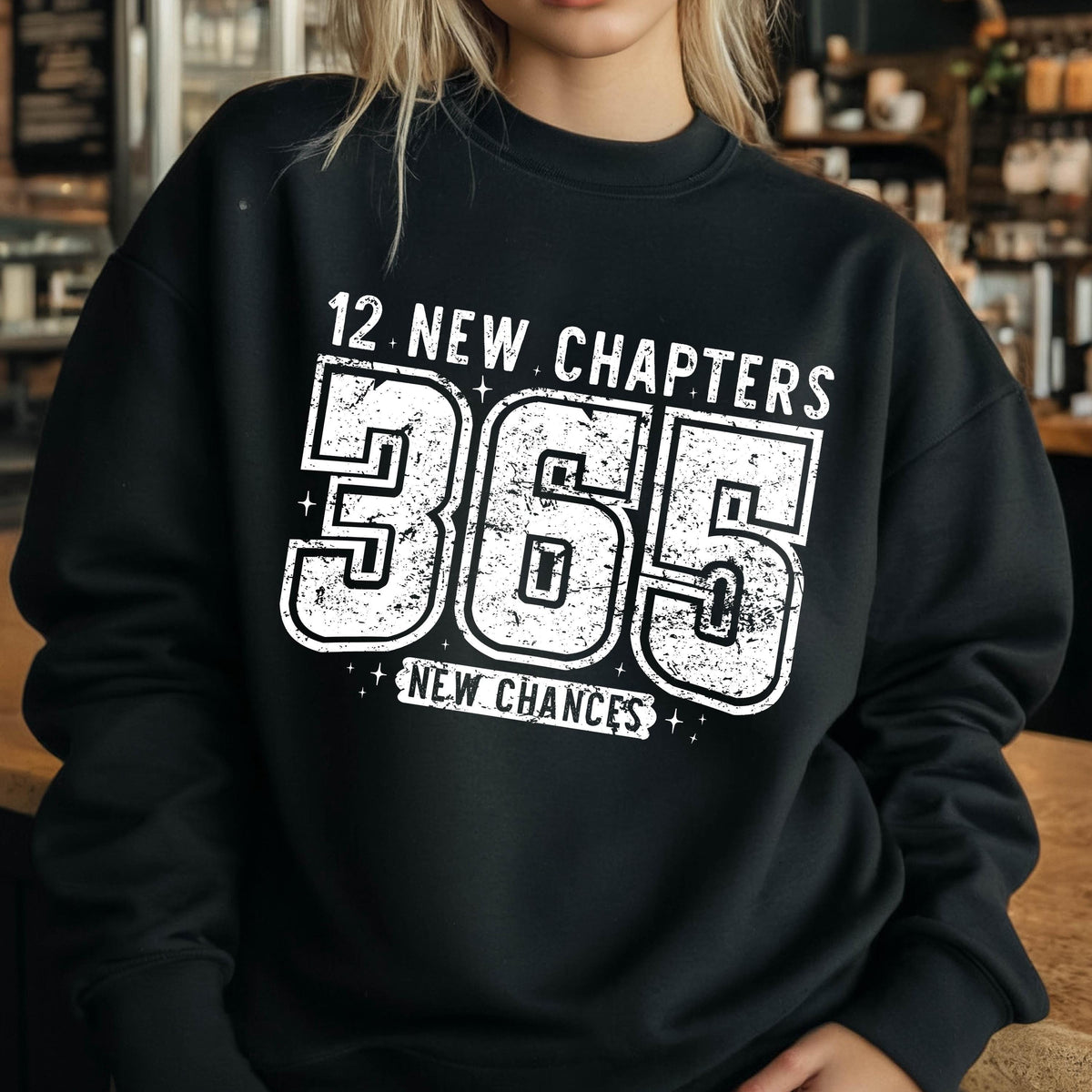 May include: Black sweatshirt with the text "12 NEW CHAPTERS 365 NEW CHANCES" in distressed white lettering. The sweatshirt is a crew neck style, perfect for casual wear. The design is centered on the front of the garment.