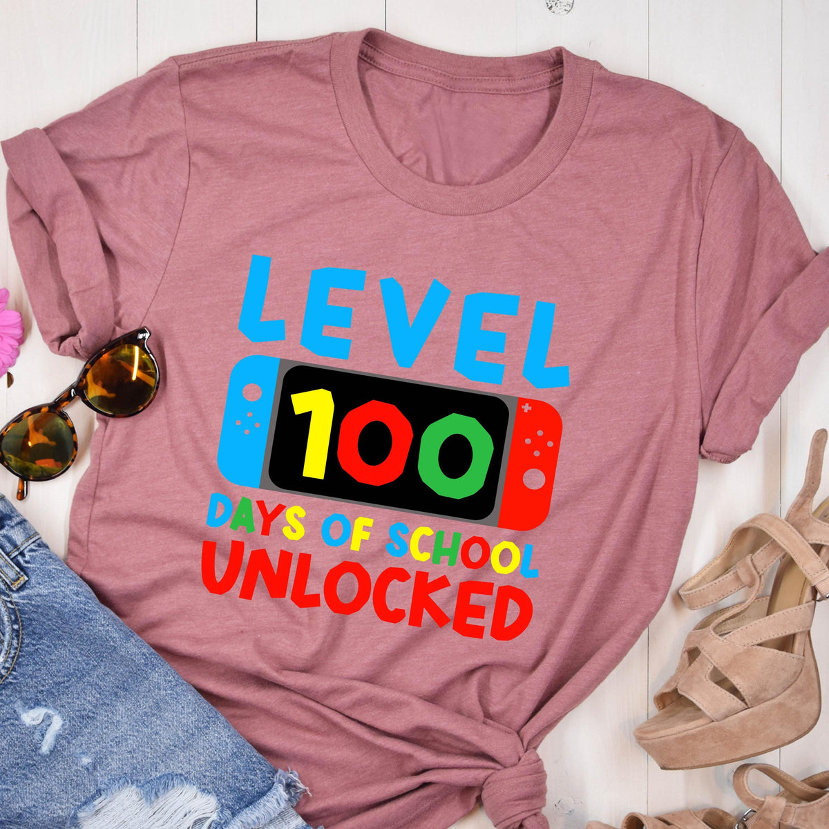 May include: A pink t-shirt with the text "LEVEL 100 DAYS OF SCHOOL UNLOCKED" in colorful letters. The text is stylized to look like a video game screen with a controller on either side.