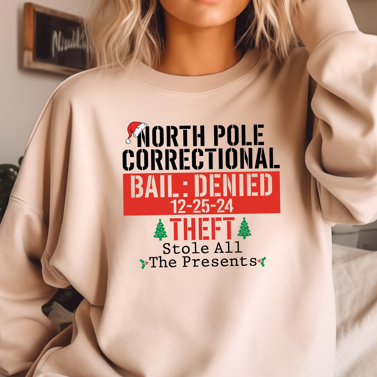 May include: A beige sweatshirt with a humorous Christmas-themed graphic. The design includes the text "NORTH POLE CORRECTIONAL BAIL: DENIED 12-25-24 THEFT Stole All The Presents" in black and red, with a Santa hat and Christmas trees.