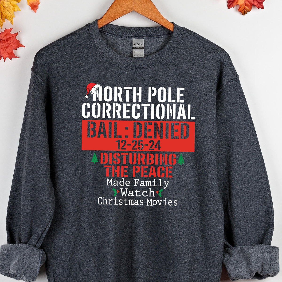 May include: Dark gray sweatshirt with the text "NORTH POLE CORRECTIONAL BAIL: DENIED 12-25-24 DISTURBING THE PEACE Made Family Watch Christmas Movies". The text is in white and red.