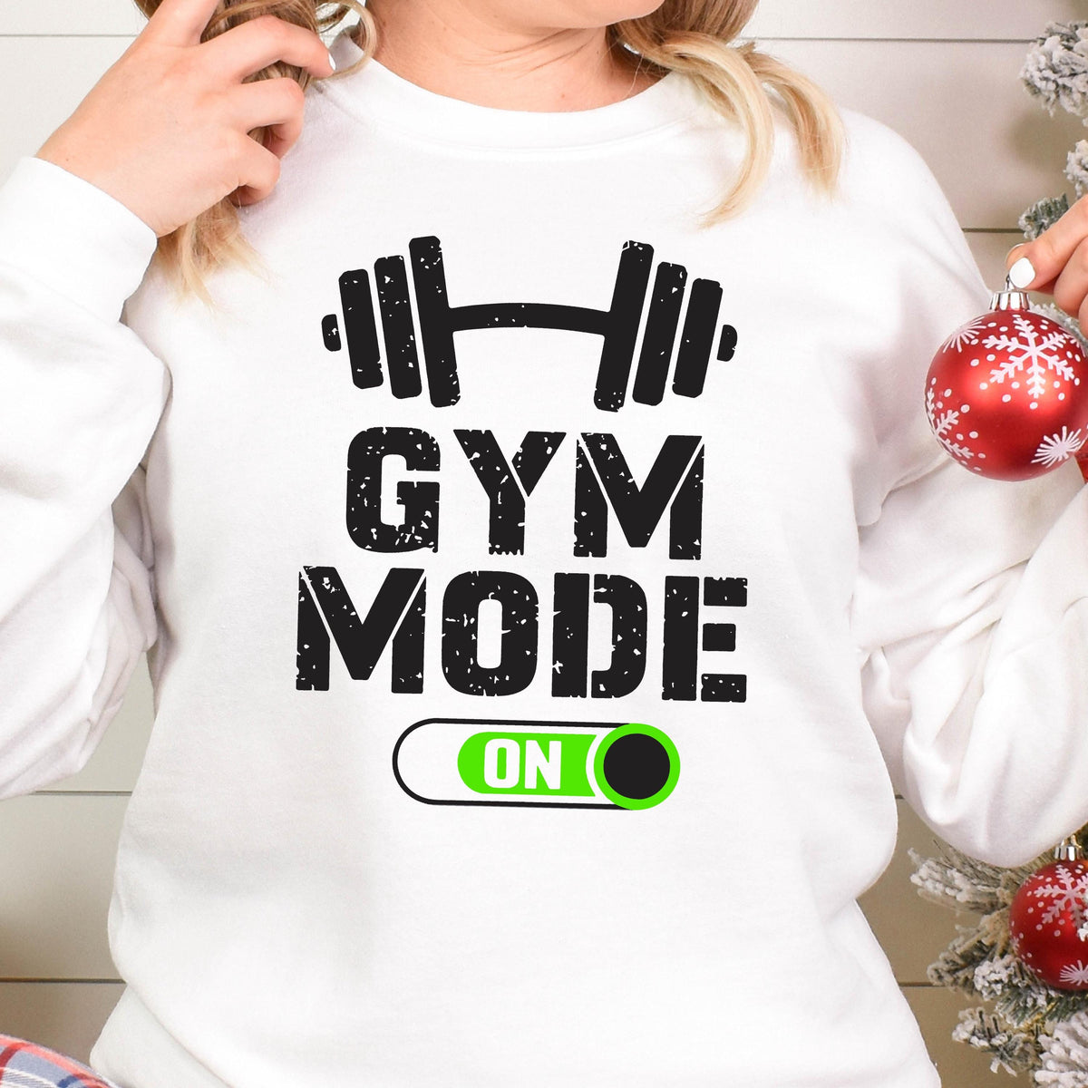 May include: A white long-sleeve sweatshirt featuring a black graphic of a barbell above the text GYM MODE in a distressed font. Below the text is a horizontal toggle switch graphic with a bright green background and the word ON in white.