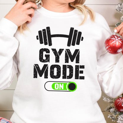 May include: A white long-sleeve sweatshirt featuring a black graphic of a barbell above the text GYM MODE in a distressed font. Below the text is a horizontal toggle switch graphic with a bright green background and the word ON in white.