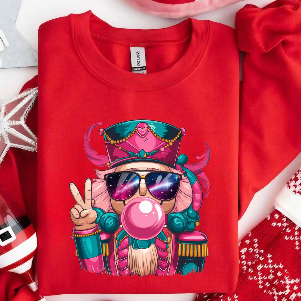 May include: A red crewneck sweatshirt featuring a cartoon nutcracker wearing sunglasses and blowing a bubblegum bubble. The nutcracker is wearing a hat and giving a peace sign. The design includes pink, teal, and gold accents.