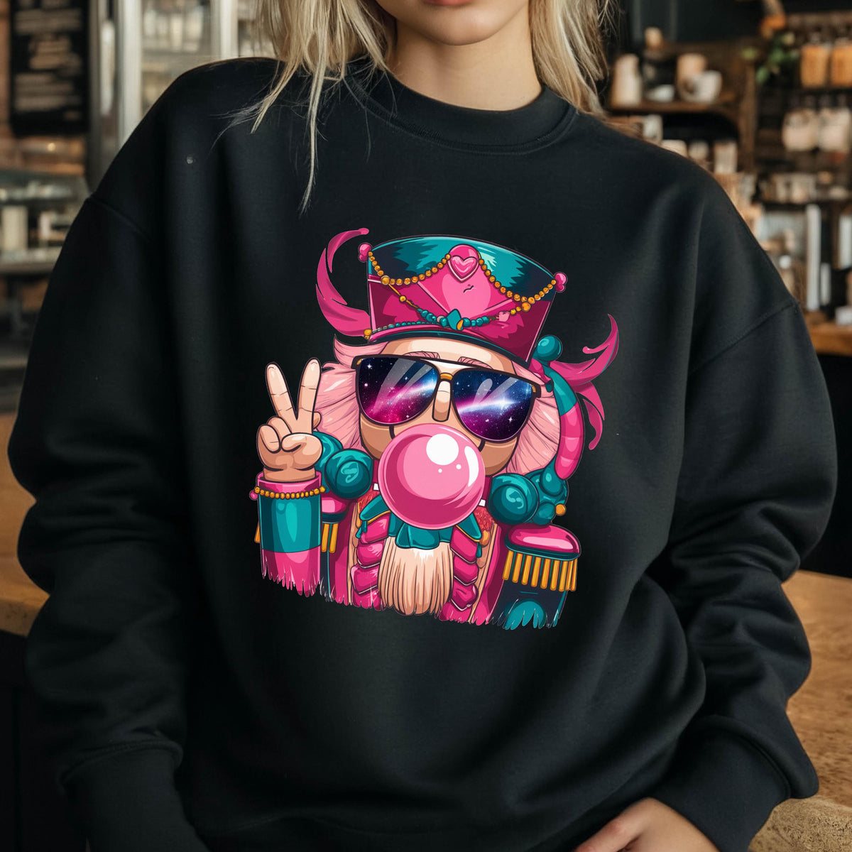 May include: Black sweatshirt featuring a colorful illustration of a nutcracker wearing sunglasses and a pink hat. The nutcracker is blowing a bubblegum bubble and making a peace sign with its hand. The design has pink, teal, and gold accents.