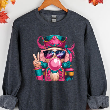 May include: Dark gray sweatshirt featuring a colorful graphic of a nutcracker wearing sunglasses and blowing a bubblegum bubble. The nutcracker is wearing a pink and teal hat and uniform, and is giving a peace sign.