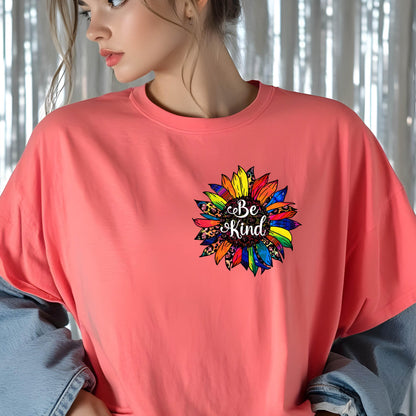 May include: A coral-colored t-shirt featuring a colorful sunflower graphic with the words "Be Kind" in a script font. The sunflower design incorporates various patterns and colors, including leopard print and rainbow hues. The shirt has a crew neck and short sleeves.