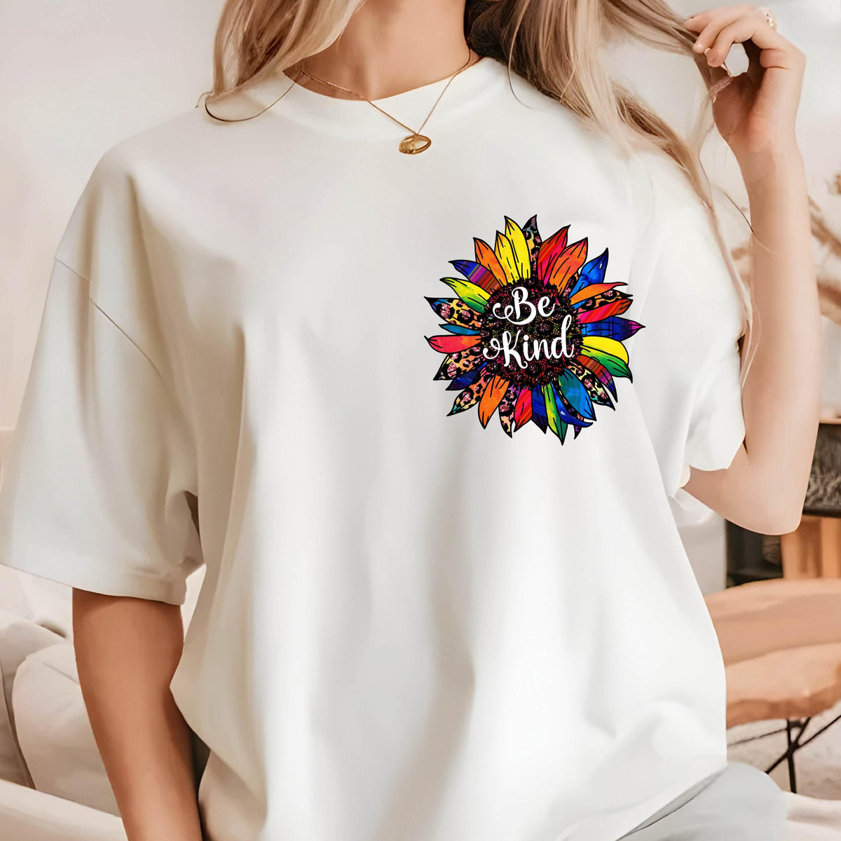 May include: White t-shirt featuring a colorful sunflower design with the words "Be Kind" in a script font. The sunflower has a rainbow of colors and a leopard print center. The shirt has short sleeves and a crew neck.