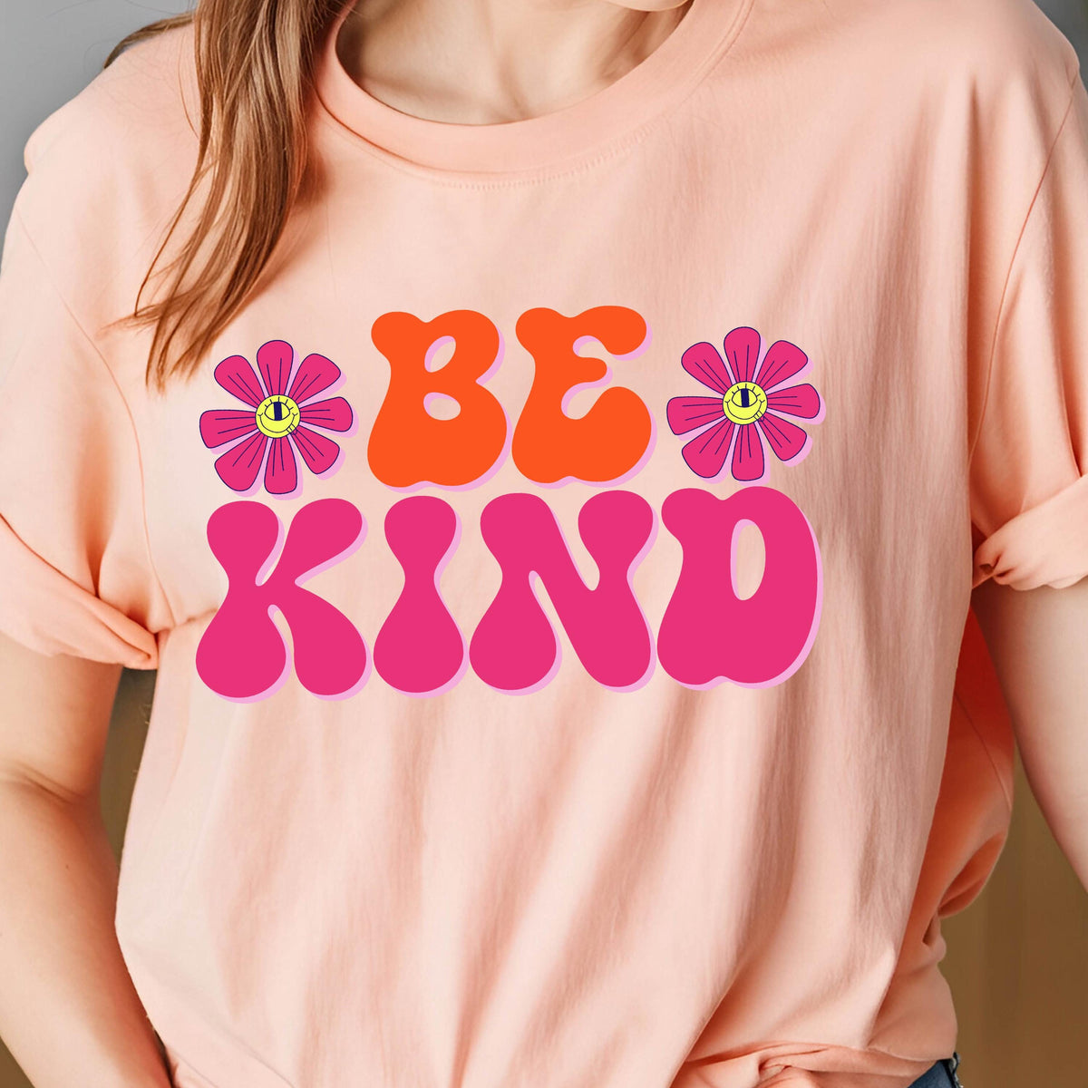 May include: A pink t-shirt with the words "BE KIND" in large, retro-style letters. The letters are pink and orange with a pink flower on each side.