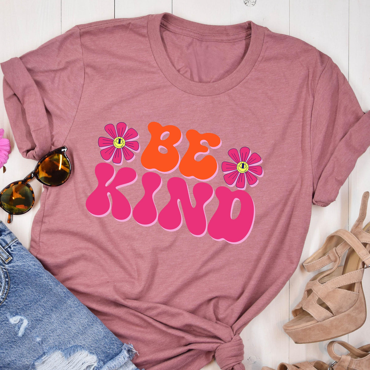 May include: A pink t-shirt with the words "BE KIND" in a retro font. The words are in orange and pink, and there are two pink flowers with yellow centers on either side of the words.