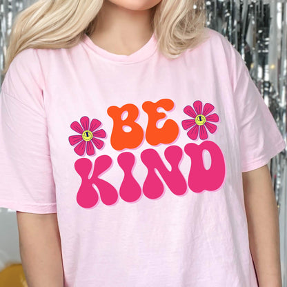 May include: Pink t-shirt with the text "BE KIND" in large, colorful, retro-style letters. The letters are outlined in pink and filled with orange. Two pink flowers with yellow smiley faces are on either side of the text.