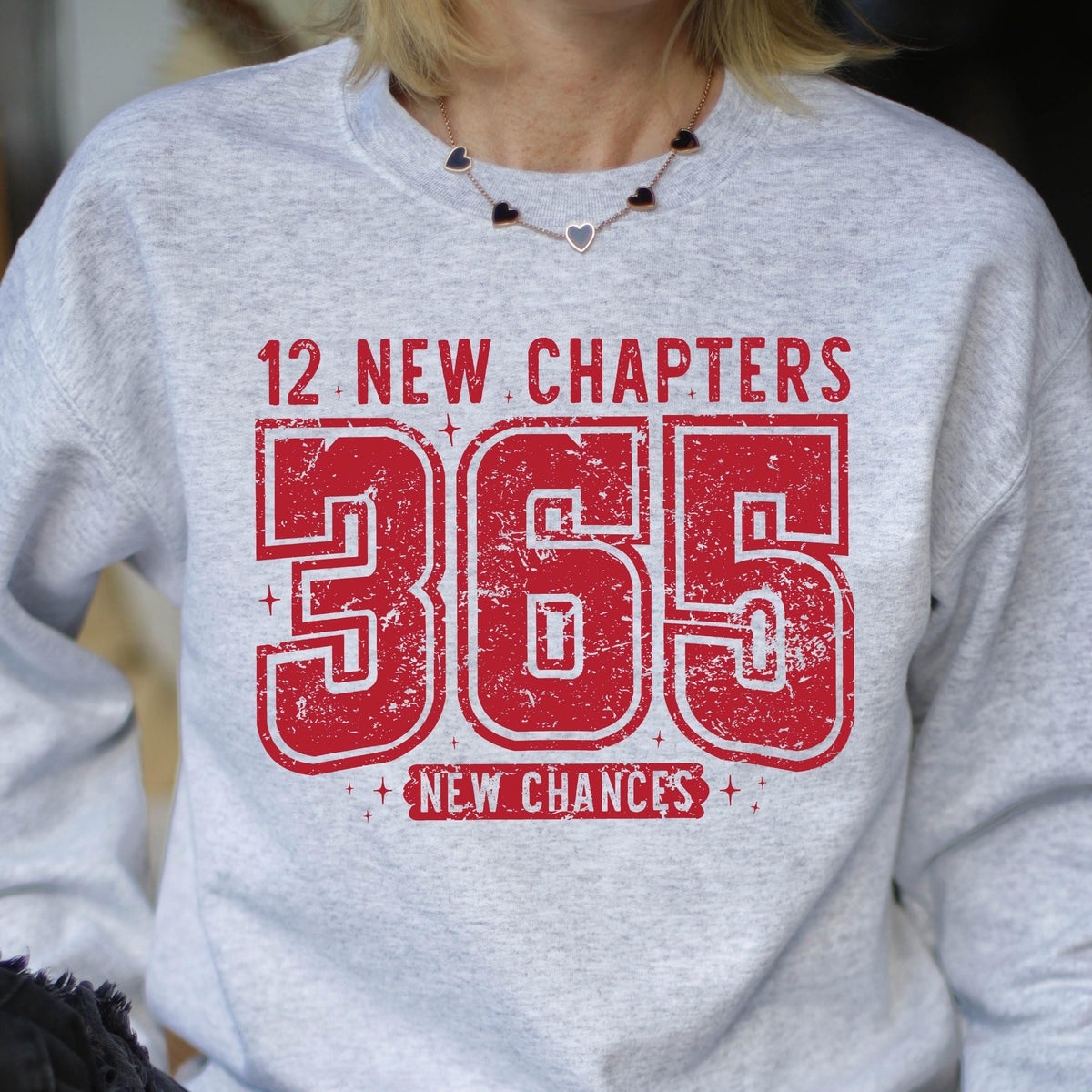 May include: A light gray sweatshirt with the text "12 NEW CHAPTERS 365 NEW CHANCES" in distressed red lettering. The sweatshirt has a crew neck and long sleeves. The text is centered on the front of the shirt.