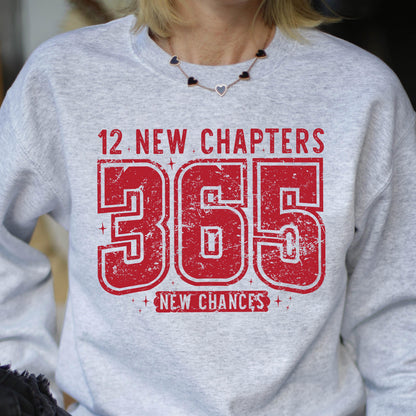 May include: A light gray sweatshirt with the text "12 NEW CHAPTERS 365 NEW CHANCES" in distressed red lettering. The sweatshirt has a crew neck and long sleeves. The text is centered on the front of the shirt.