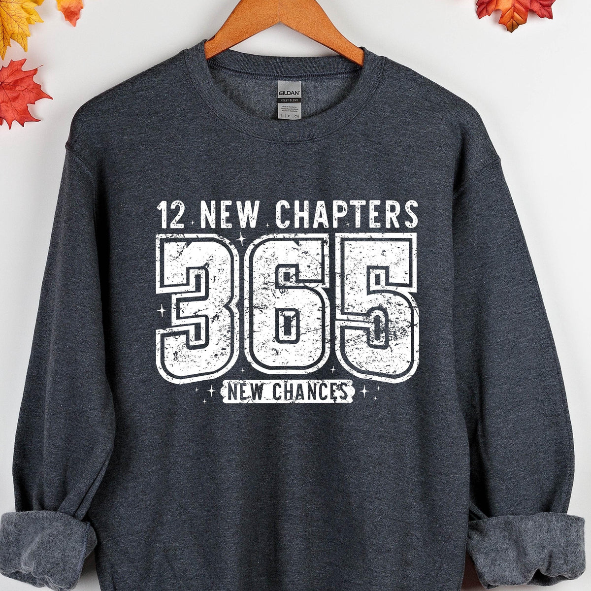 May include: Dark gray sweatshirt with the text "12 NEW CHAPTERS 365 NEW CHANCES" in white. The sweatshirt is hanging on a wooden hanger. The text is in a bold, blocky font.