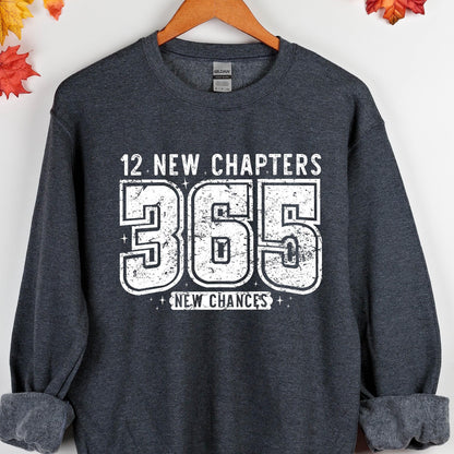 May include: Dark gray sweatshirt with the text "12 NEW CHAPTERS 365 NEW CHANCES" in white. The sweatshirt is hanging on a wooden hanger. The text is in a bold, blocky font.