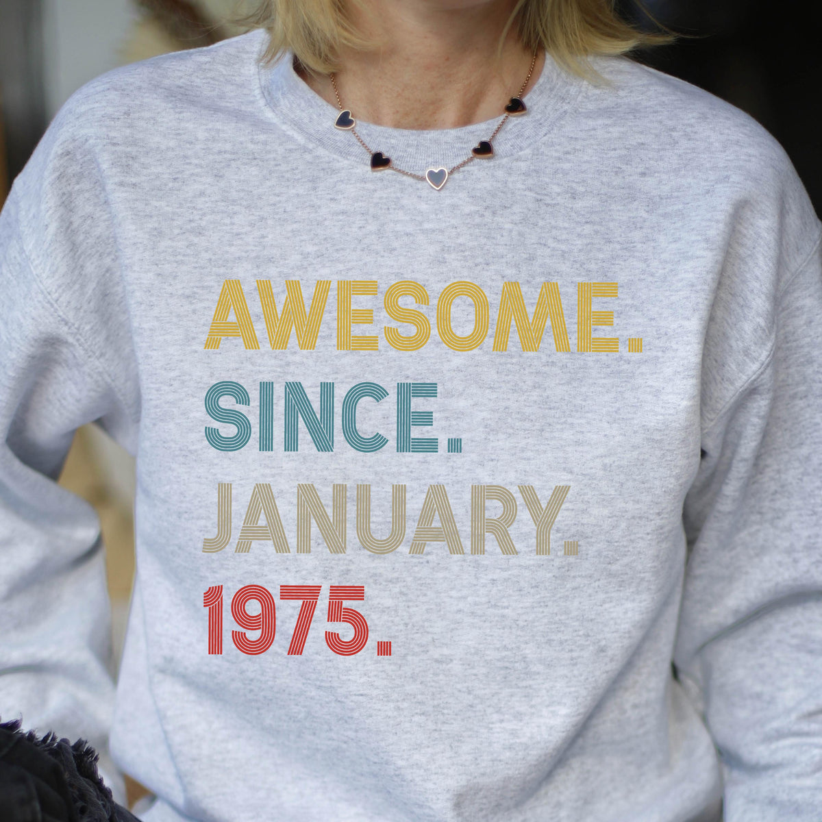 May include: A light gray sweatshirt with the text "AWESOME. SINCE. JANUARY. 1975." in yellow, teal, tan, and red. The sweatshirt is a crew neck style.