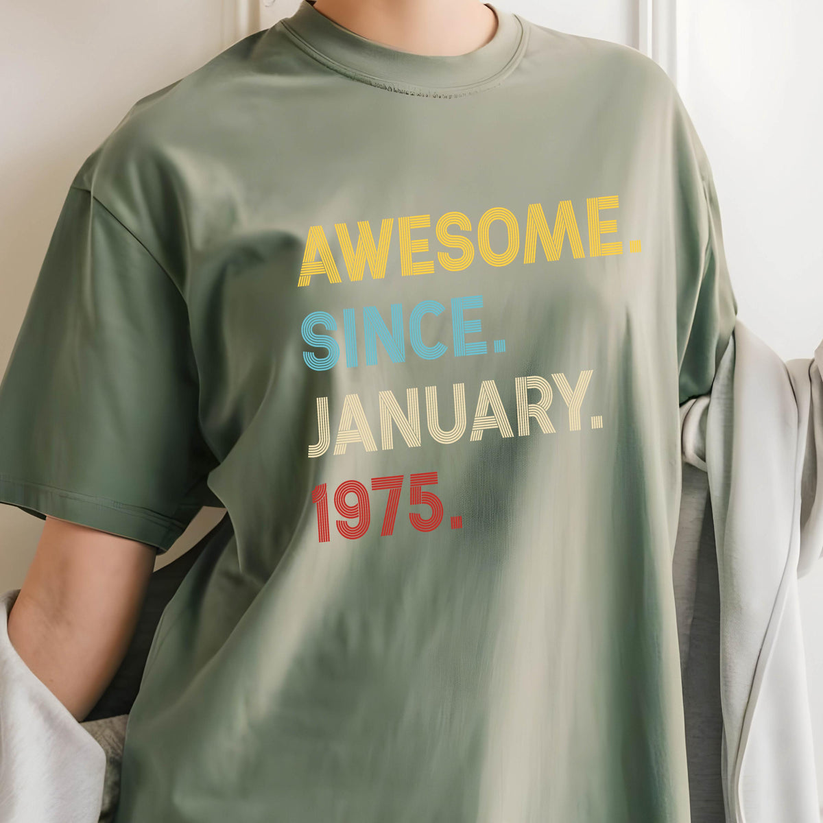 May include: An olive green crew neck t-shirt featuring the text AWESOME. SINCE. JANUARY. 1975. printed in a stacked design. The word AWESOME is yellow, SINCE is light blue, JANUARY is cream, and 1975 is red, all with a subtle line texture.