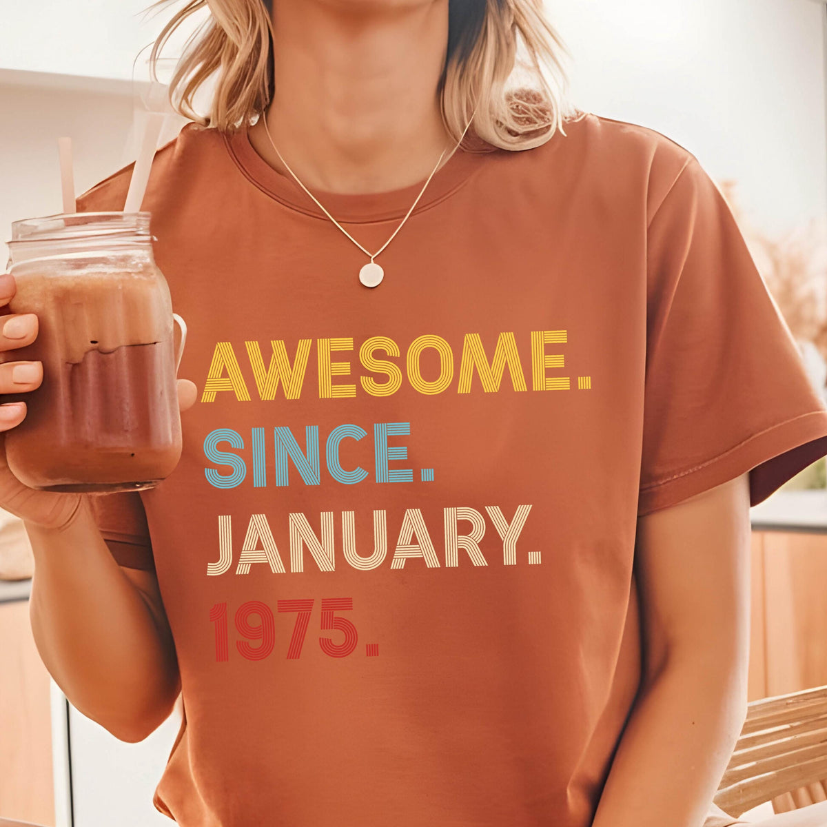 May include: A rust-colored short-sleeve t-shirt featuring the text AWESOME. SINCE. JANUARY. 1975. printed in yellow, light blue, white, and red. The shirt has a crew neckline and a straight hem, with the text displayed in a multi-line graphic design.