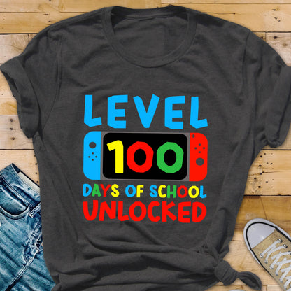 May include: A dark gray t-shirt with the text "LEVEL 100 DAYS OF SCHOOL UNLOCKED" in colorful letters. The text is stylized to look like a video game screen with a controller on either side.