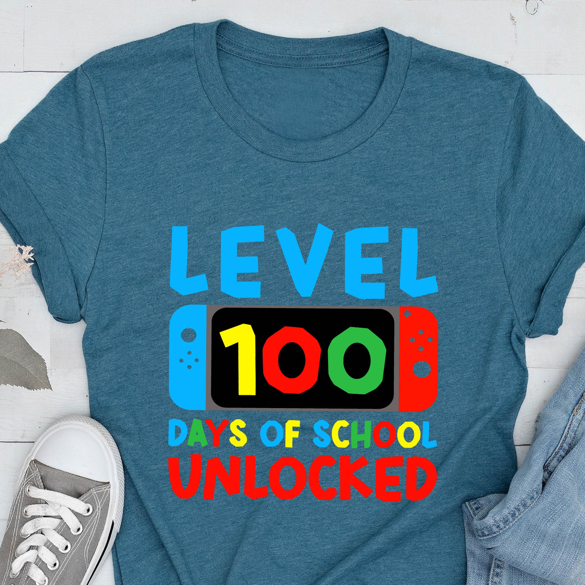 May include: A teal blue t-shirt with the text "Level 100 Days of School Unlocked" in colorful letters. The text is displayed on a graphic of a video game controller with a screen showing the number 100.