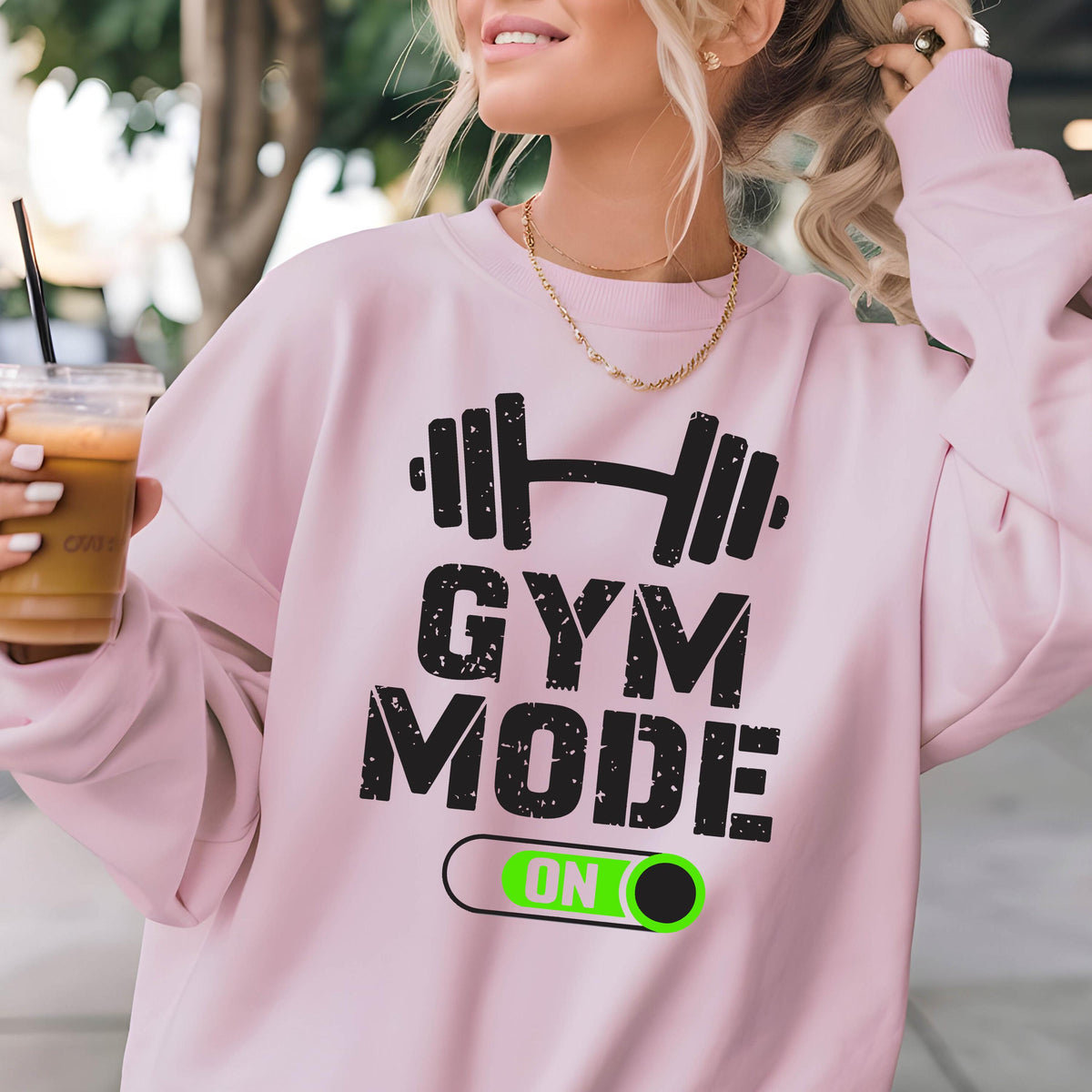 May include: A light pink long-sleeve sweatshirt featuring a black graphic of a barbell above the text GYM MODE and a toggle switch graphic with the word ON in bright green. The sweatshirt has a crew neckline and a relaxed fit design.