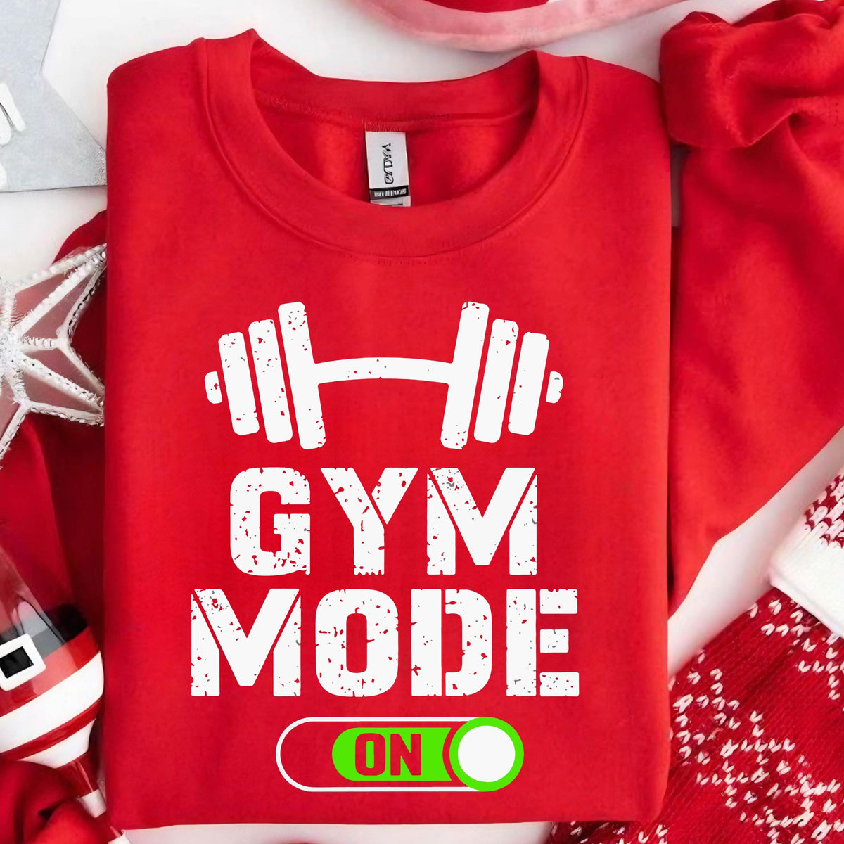 May include: A bright red crewneck sweatshirt featuring a white graphic of a barbell above the text GYM MODE and a green toggle switch graphic with the word ON. The design has a distressed, weathered texture throughout the white lettering.