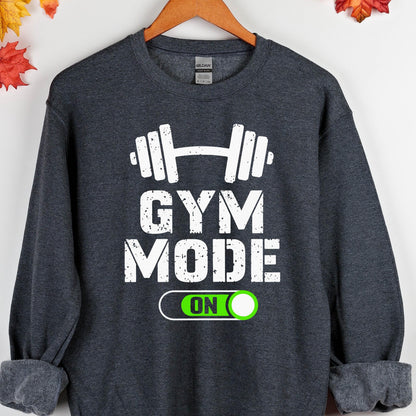 May include: A dark heather gray crewneck sweatshirt featuring a white graphic of a barbell above the text GYM MODE in a distressed font. Below the text is a white outlined toggle switch graphic with the word ON in a bright green circle.