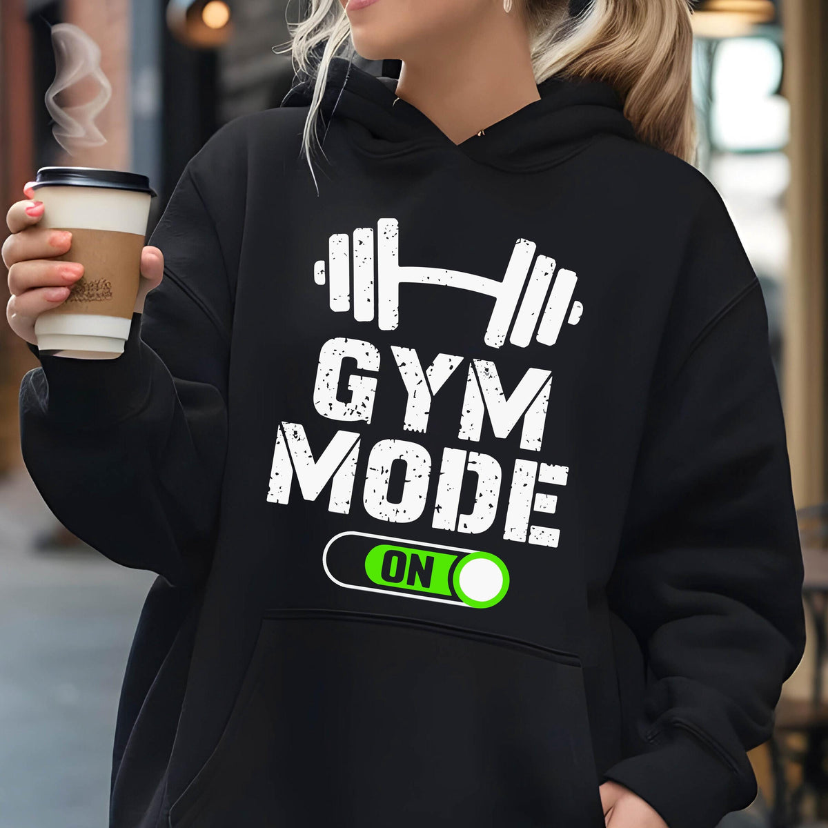 May include: A black hooded sweatshirt featuring a white graphic of a barbell above the text GYM MODE and a green toggle switch graphic with the word ON. The design has a distressed texture and the hoodie includes a front kangaroo pocket.