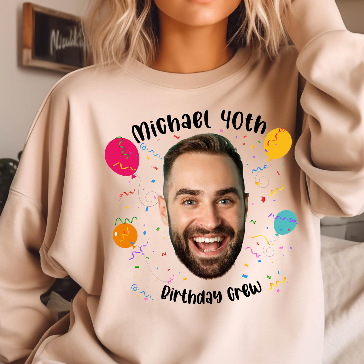 May include: A light pink sweatshirt with a black graphic design featuring a man's face with a beard, surrounded by colorful balloons and confetti. The text "Michael 40th Birthday Crew" is printed on the sweatshirt.