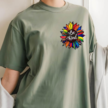 May include: Sage green t-shirt featuring a colorful sunflower design with the words "Be Kind" in a script font. The sunflower has a mix of rainbow and leopard print petals. The shirt has short sleeves and a crew neck.