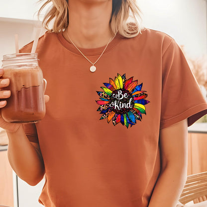 May include: A rust-colored t-shirt featuring a colorful sunflower design with the words "Be Kind" in the center. The sunflower has petals in various colors, including yellow, blue, and red. The shirt is made of a soft material.