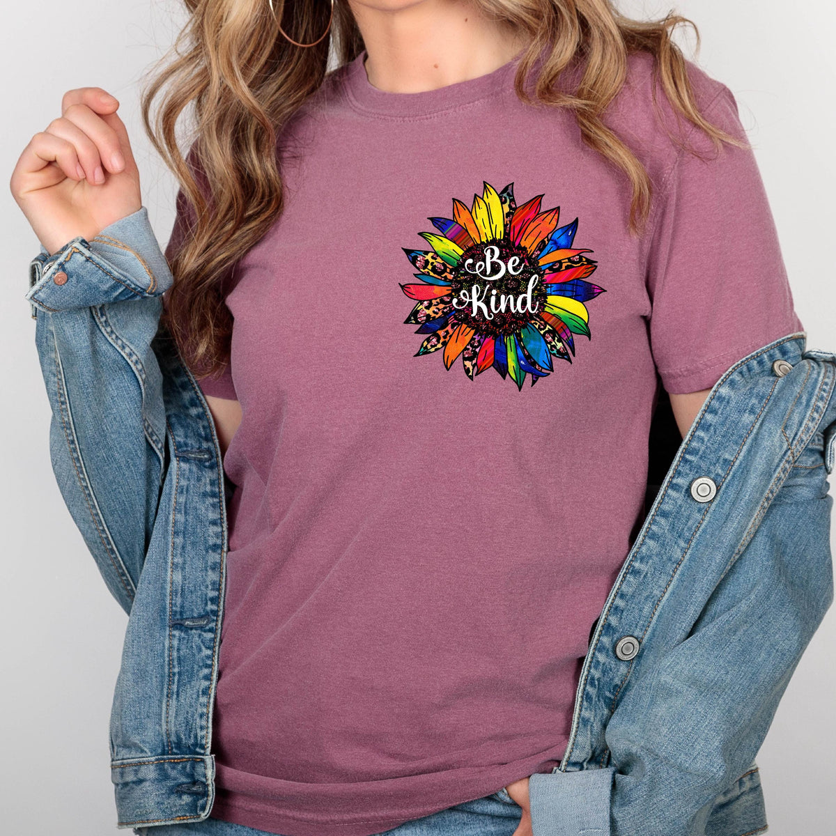 May include: Mauve short-sleeved t-shirt with a colorful sunflower graphic on the chest. The sunflower design features rainbow petals, leopard print accents, and the words "Be Kind" in a script font.