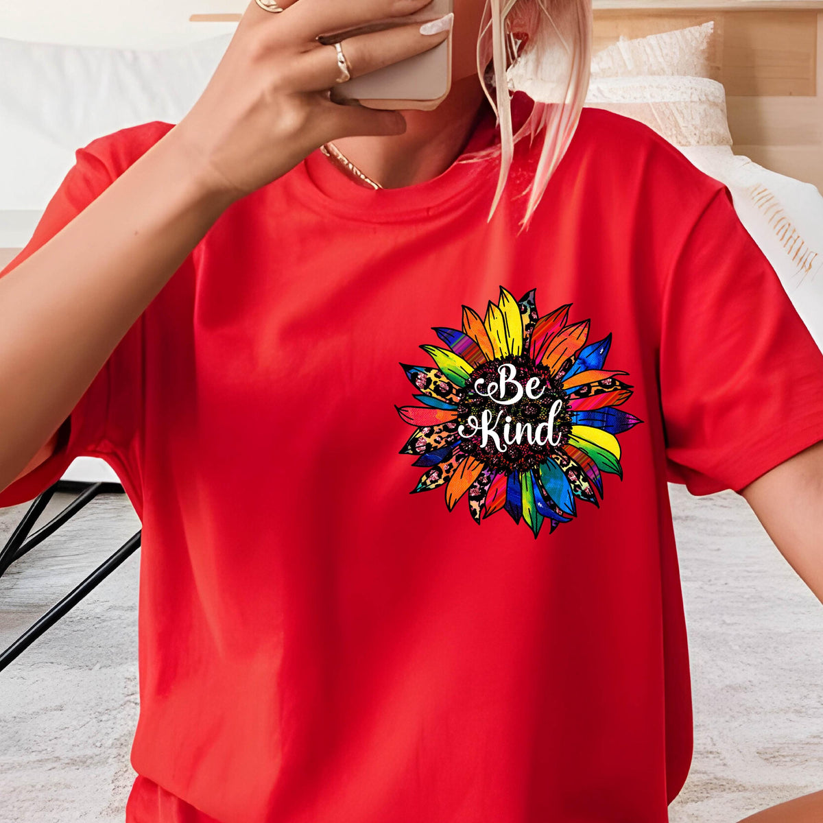 May include: A red t-shirt featuring a colorful sunflower design with the words "Be Kind" in a script font. The sunflower has yellow, blue, green, and leopard print petals. The shirt is short-sleeved.