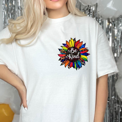 May include: White t-shirt featuring a colorful sunflower design with the words "Be Kind" in a script font. The sunflower has petals in shades of yellow, orange, red, blue, and green. The shirt is a casual, everyday wear item.