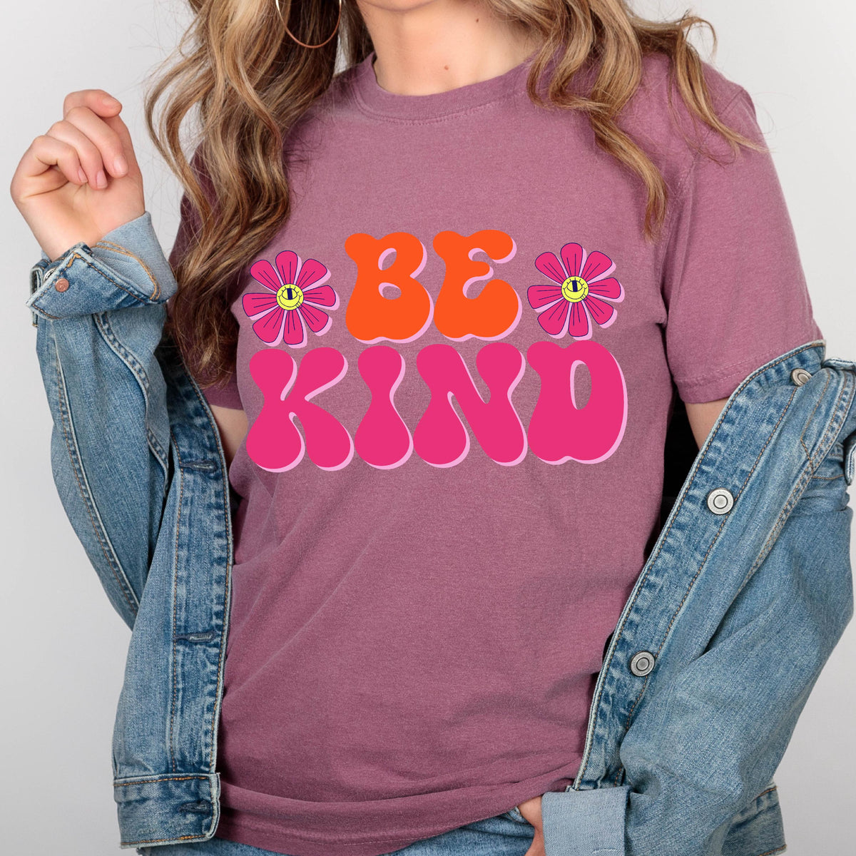 May include: A mauve t-shirt with a retro-style graphic that says "Be Kind" in pink and orange letters with two pink flowers with yellow smiley faces.