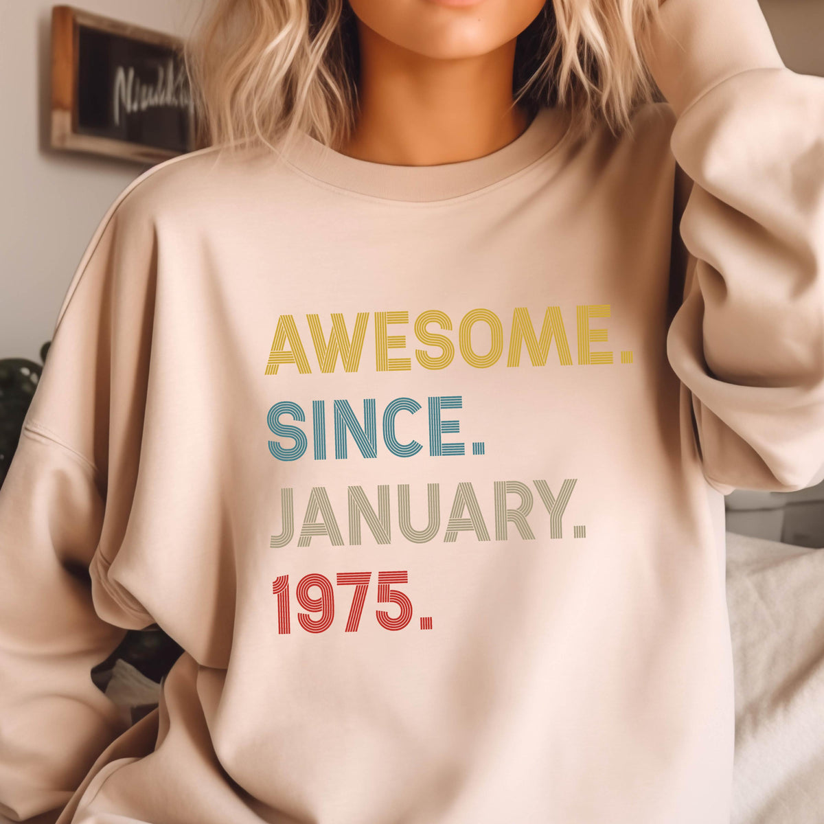 May include: Beige sweatshirt with the text "AWESOME. SINCE. JANUARY. 1975." in yellow, blue, gray, and red. The sweatshirt is a neutral color and has long sleeves.