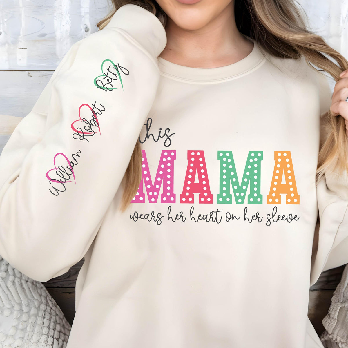 May include: A cream-colored sweatshirt with the text "This mama wears her heart on her sleeve" in colorful letters with white polka dots. The sleeves have the names "William, Robert, Betty" written in cursive with pink, red, and green hearts.