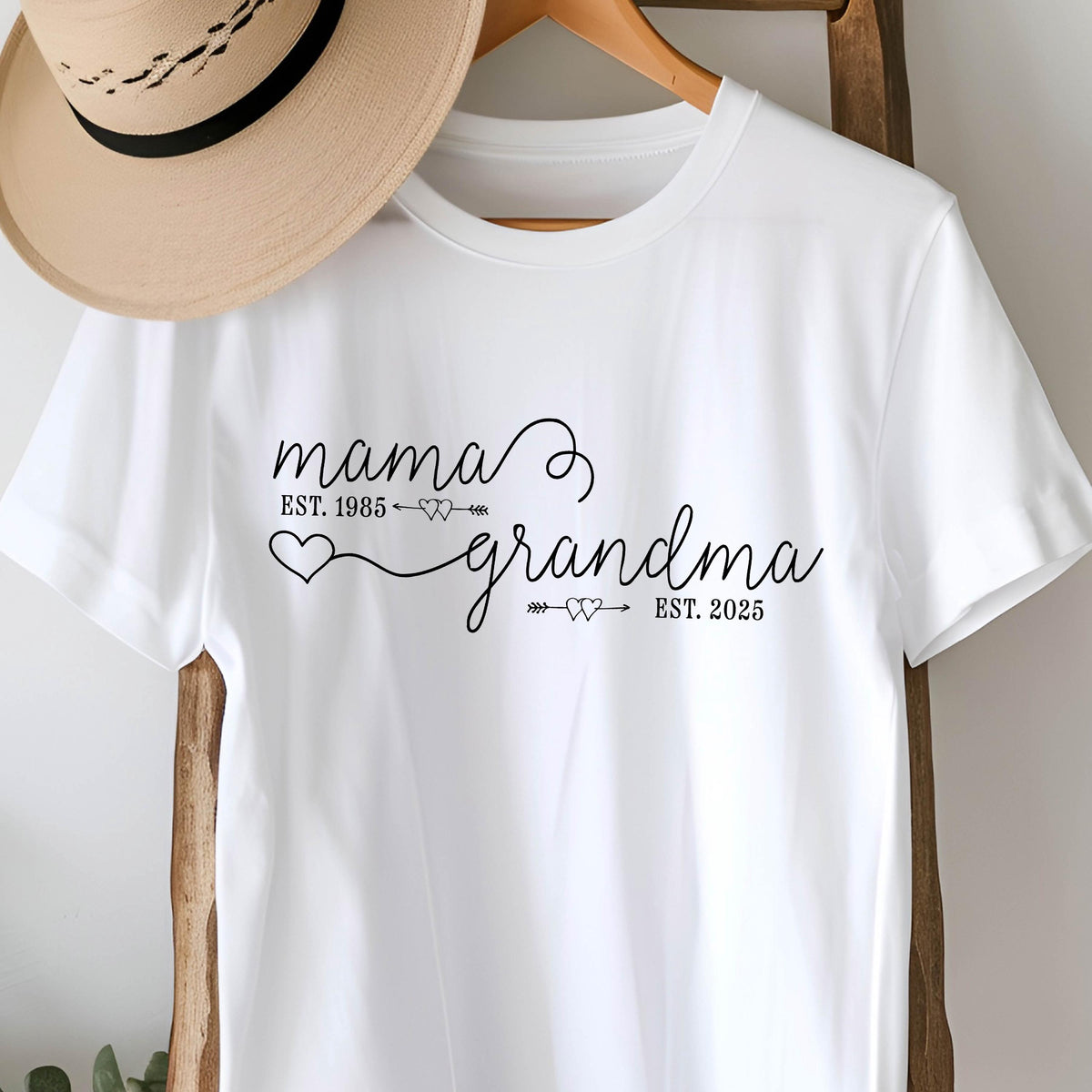 May include: White t-shirt with black text that reads "mama est. 1985" with an arrow pointing to the right and two small hearts. The text continues with "grandma est. 2025" with an arrow pointing to the left and two small hearts.