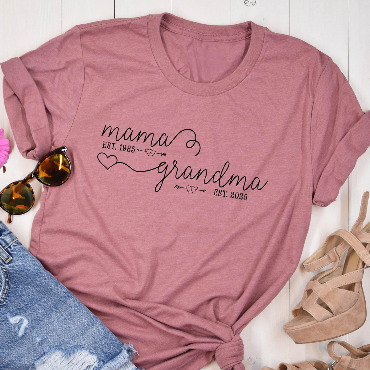 May include: A pink t-shirt with the text "mama est. 1985 grandma est. 2025" in black lettering. The text is written in a cursive font with a heart and arrow design.