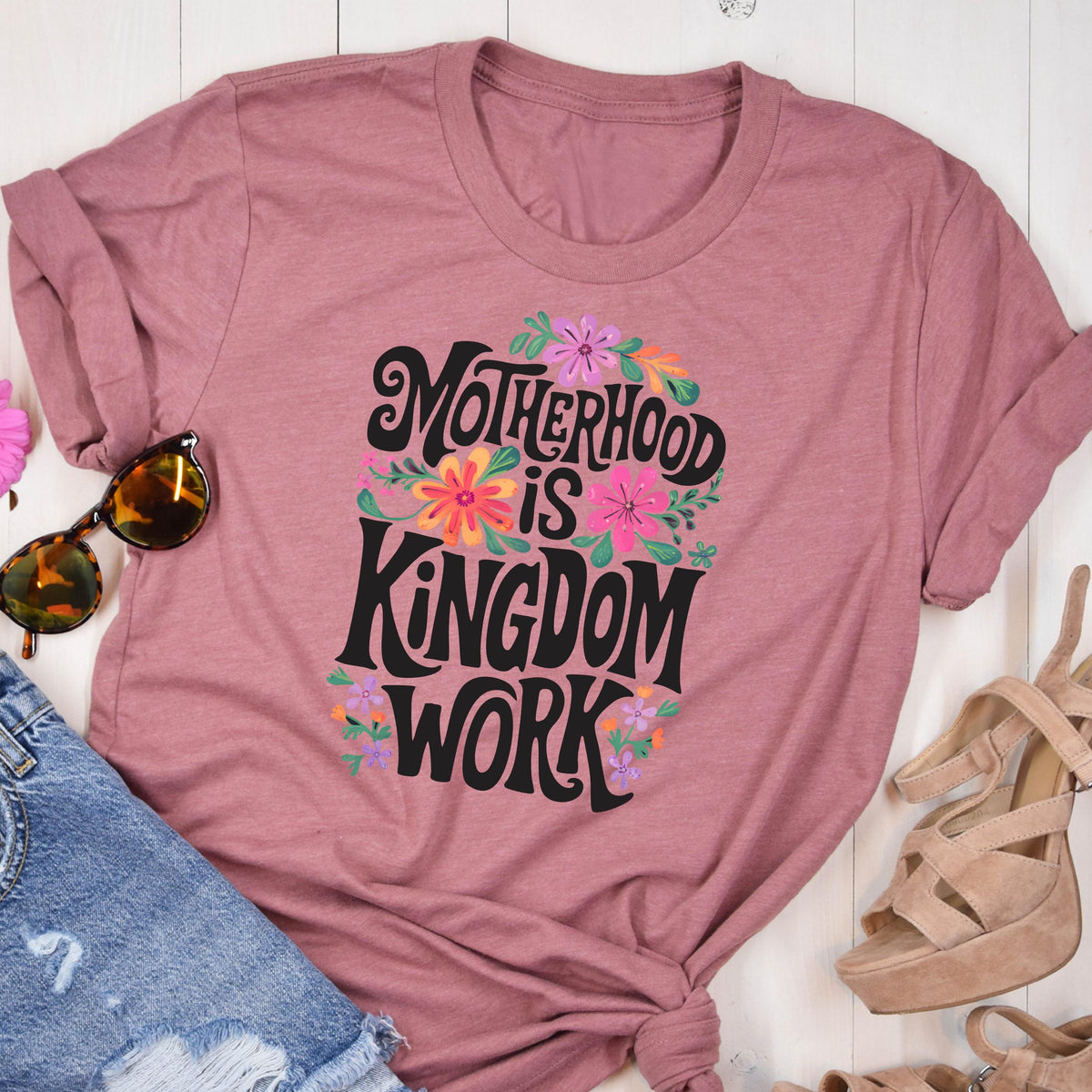 May include: A mauve t-shirt with a floral design and the text "Motherhood is Kingdom Work" in black lettering.