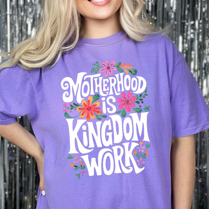 May include: Purple t-shirt with a white graphic that says "Motherhood is Kingdom Work" surrounded by colorful flowers.