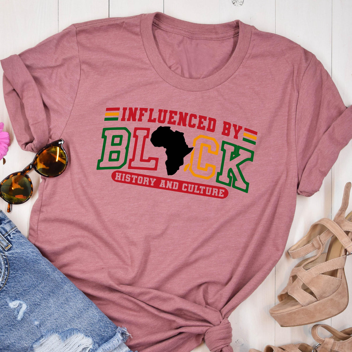 May include: A mauve short-sleeved t-shirt featuring the text "INFLUENCED BY" in red, "HISTORY AND CULTURE" in red, and the word "BLACK" in large green and yellow block letters, with a black silhouette of the African continent replacing the letter A.