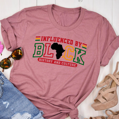 May include: A mauve short-sleeved t-shirt featuring the text "INFLUENCED BY" in red, "HISTORY AND CULTURE" in red, and the word "BLACK" in large green and yellow block letters, with a black silhouette of the African continent replacing the letter A.