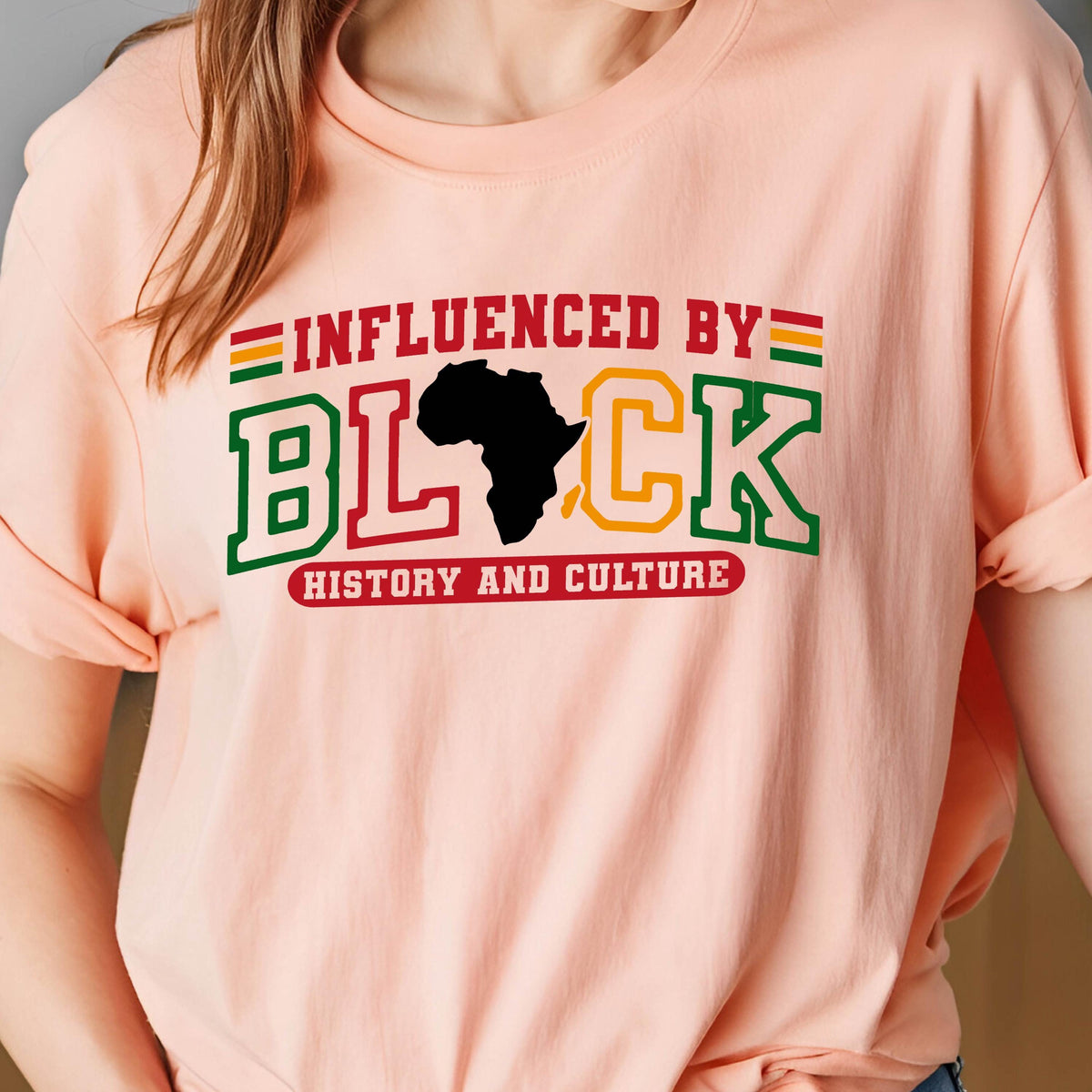 May include: A peach-colored t-shirt featuring the text INFLUENCED BY BLACK HISTORY AND CULTURE. The word BLACK is stylized with a black silhouette of the African continent replacing the letter A, with red, green, and yellow accents throughout.