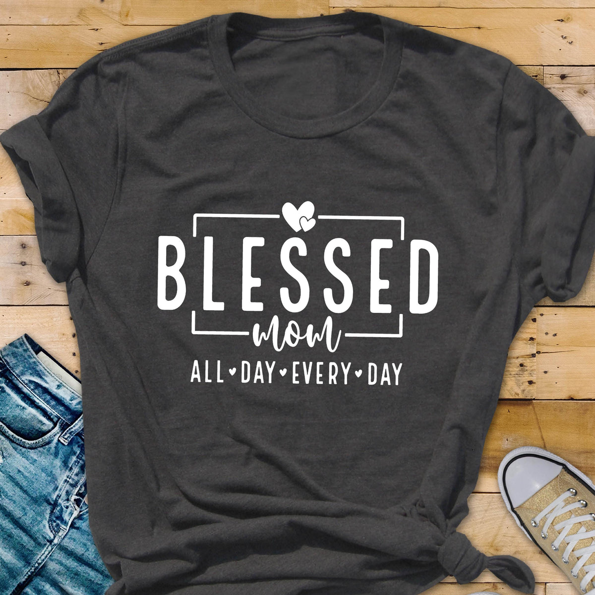 May include: A dark gray t-shirt with the text "Blessed Mom All Day Every Day" in white lettering. The text is framed by a white rectangle and includes two white hearts above the word "Mom".