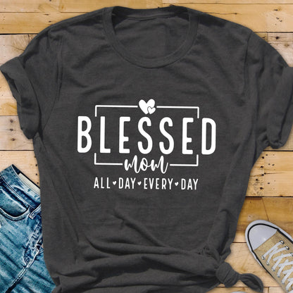 May include: A dark gray t-shirt with the text "Blessed Mom All Day Every Day" in white lettering. The text is framed by a white rectangle and includes two white hearts above the word "Mom".