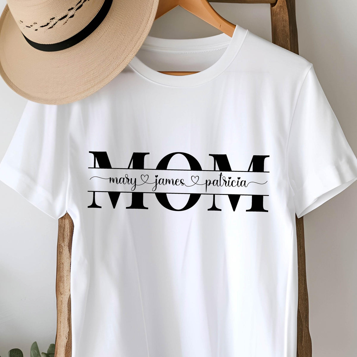May include: White t-shirt with the word "MOM" in black letters. Underneath the word "MOM" is a black line with the names "mary", "james", and "patricia" written in cursive with a heart symbol between each name.